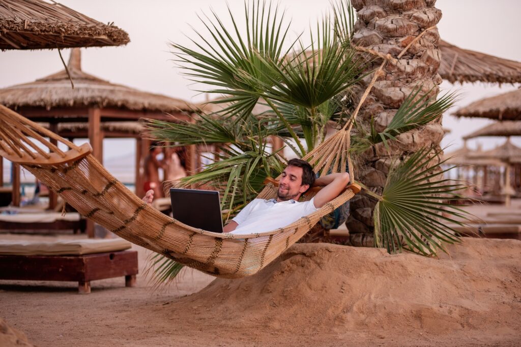 A freelance businessman lies in a hammock under palm trees, holds a laptop in his hands, works remot